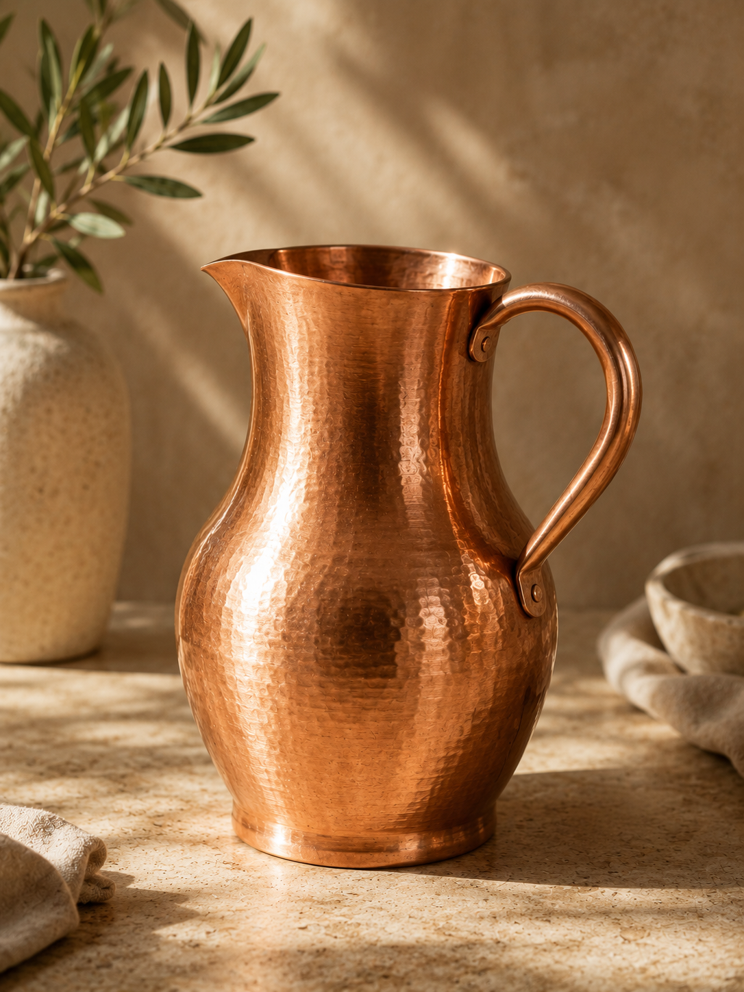 The Ambai Jug, hand-hammered copper carafe, beside a ceramic vase with an olive branch and a stone bowl, in warm natural light.