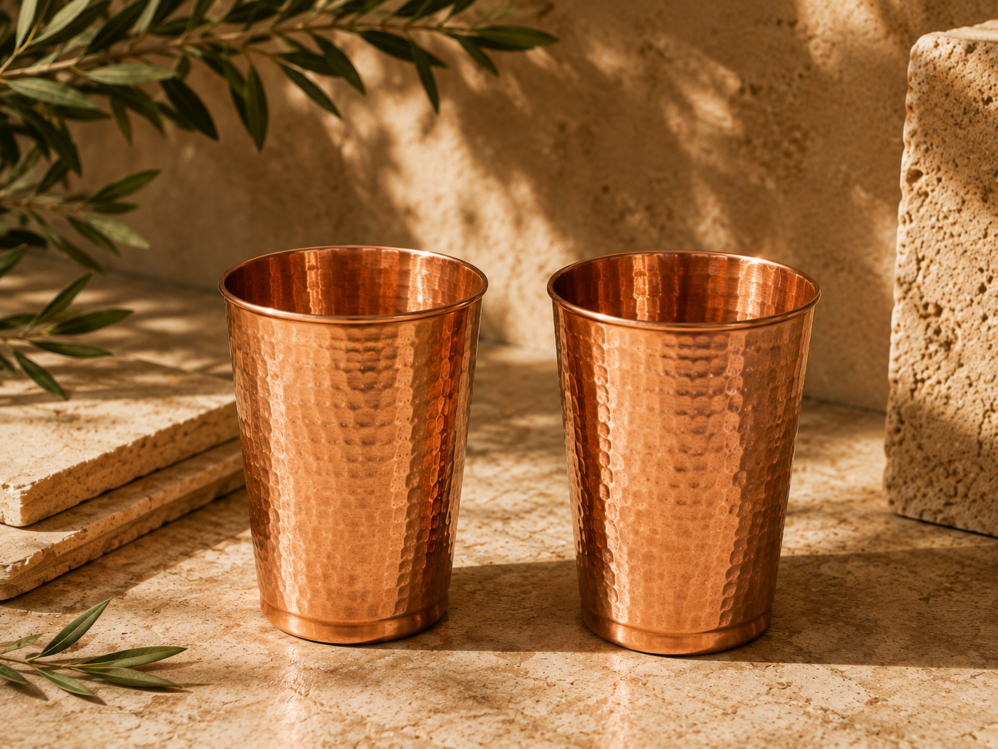 Two matching hammered copper tumblers side by side on a sunlit stone surface with an olive branch.