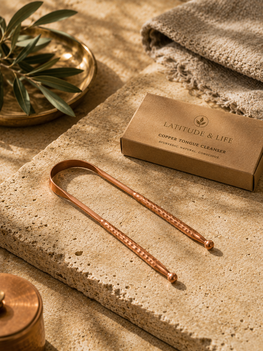 The Copper Tongue Cleanser beside its branded packaging box, on a sunlit stone surface with an olive branch.