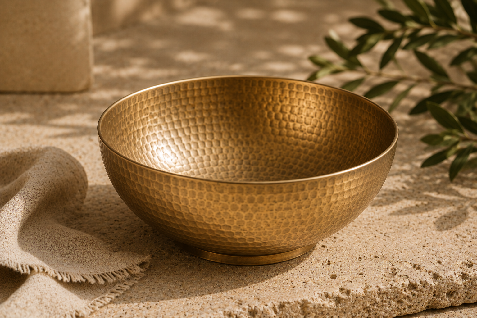 A close view of the Kansa Wellness Bowl's hammered bronze texture, on a sunlit stone surface with folded linen and an olive branch.