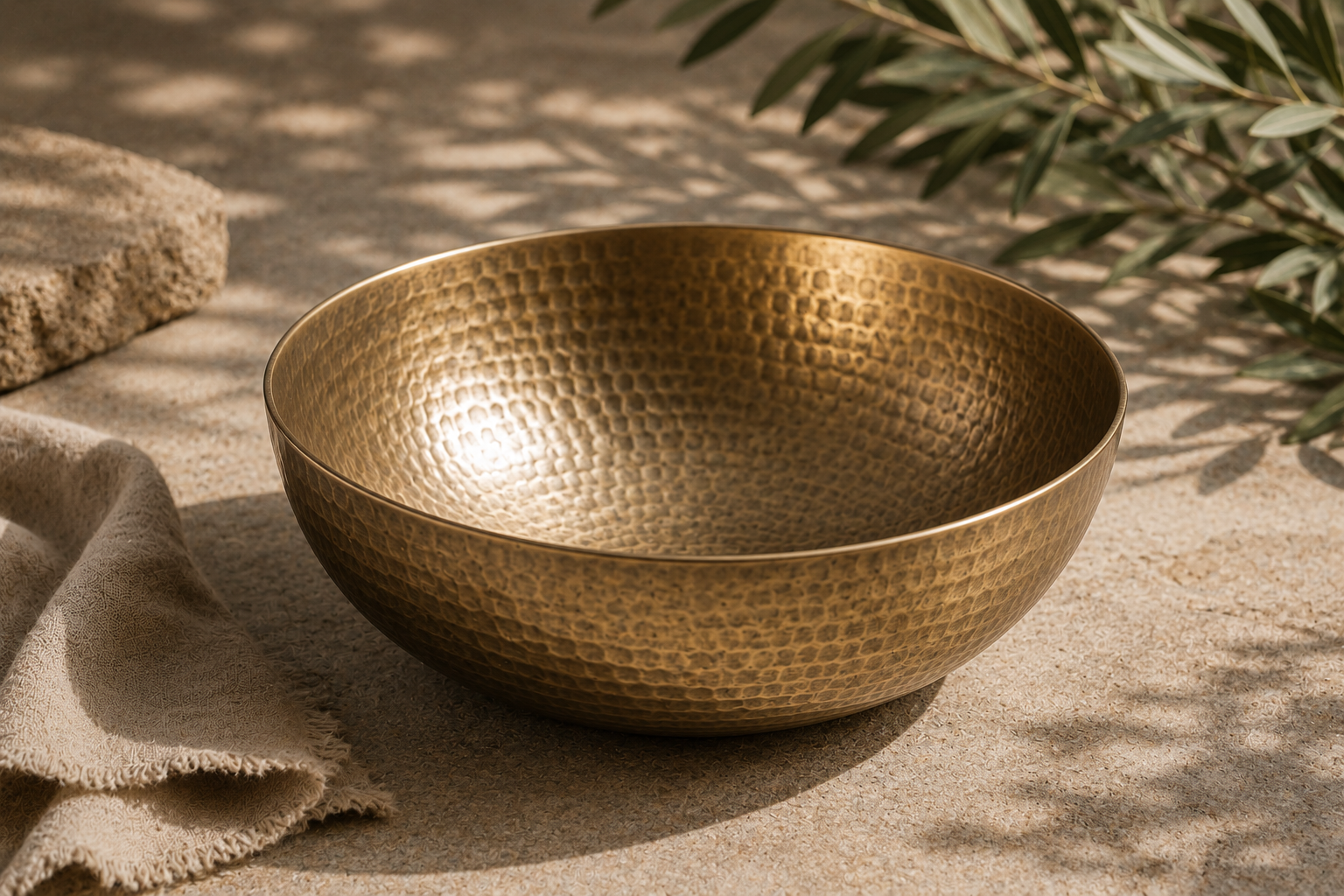 The Kansa Wellness Bowl in warm golden-bronze tone, on a sunlit stone surface with folded linen and an olive branch.