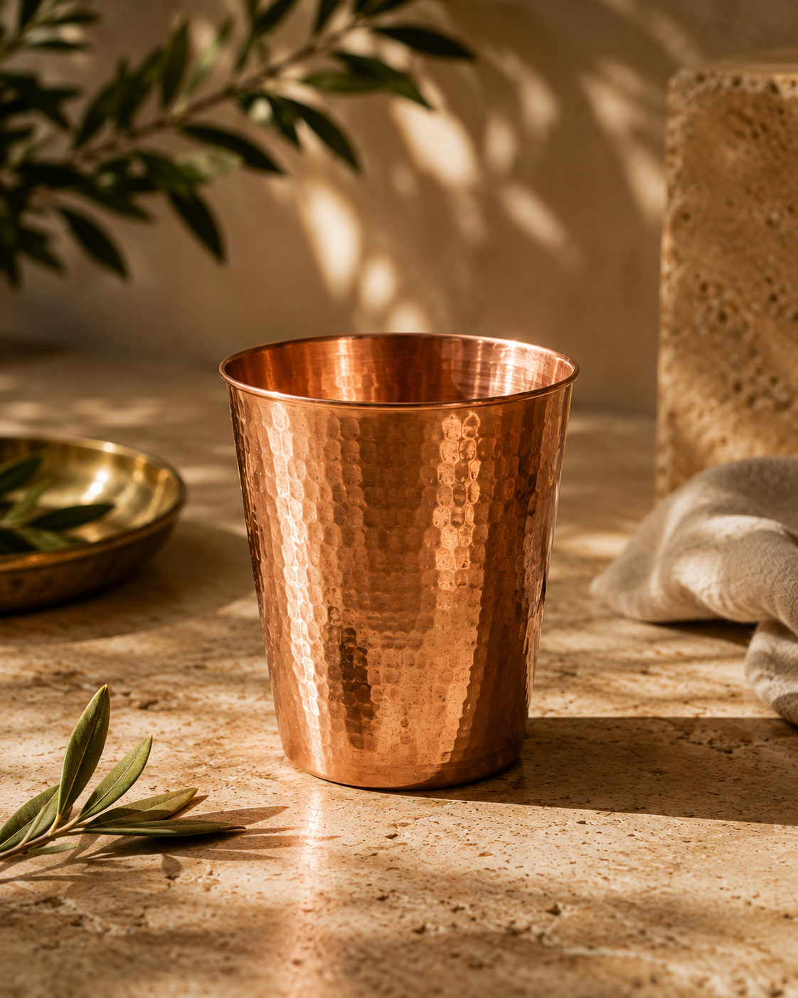 The Morning Glass, a single hammered copper tumbler, on a sunlit stone surface with an olive branch and a brass bowl.